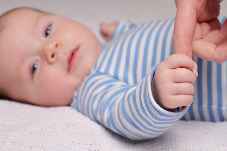 infant attachment-Baby in striped onesie grasps an adults finger and gazes into the camera