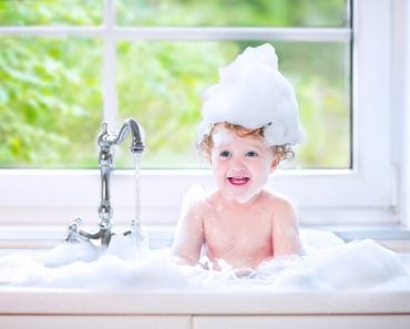 Funny little baby girl with wet curly hair taking a bath in a kitchen sink with lots of foam playing with water drops and splashes next to a big window with garden view