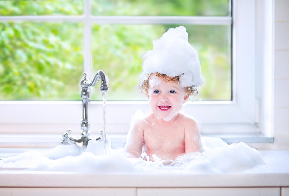 Funny little baby girl with wet curly hair taking a bath in a kitchen sink with lots of foam playing with water drops and splashes next to a big window with garden view