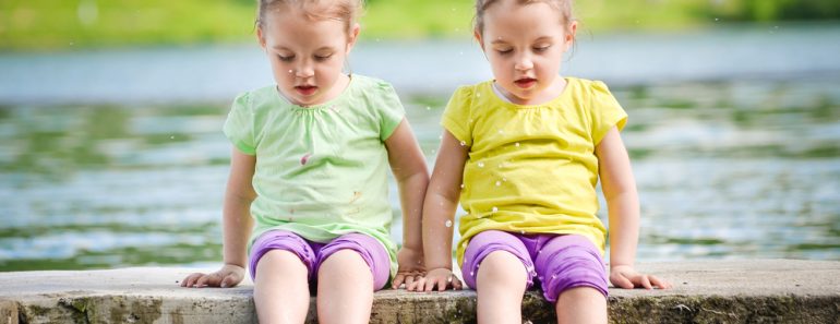 Identical twins girls are exercising on lake shore, sprinkling water. Children sitting on lake side, playing with water. Healthy and active children lifestyle. - Image(JGA)s
