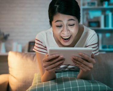 happy teenage asian girl sitting on couch at home enjoy funny movie on tablet in dark living room(PR Image Factory)s