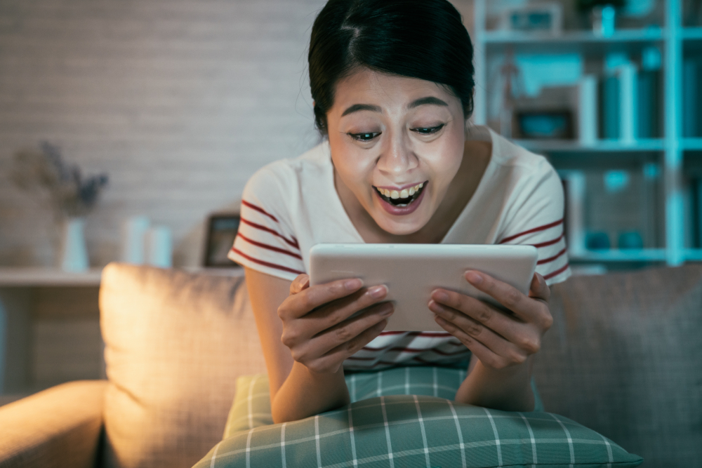 happy teenage asian girl sitting on couch at home enjoy funny movie on tablet in dark living room(PR Image Factory)s