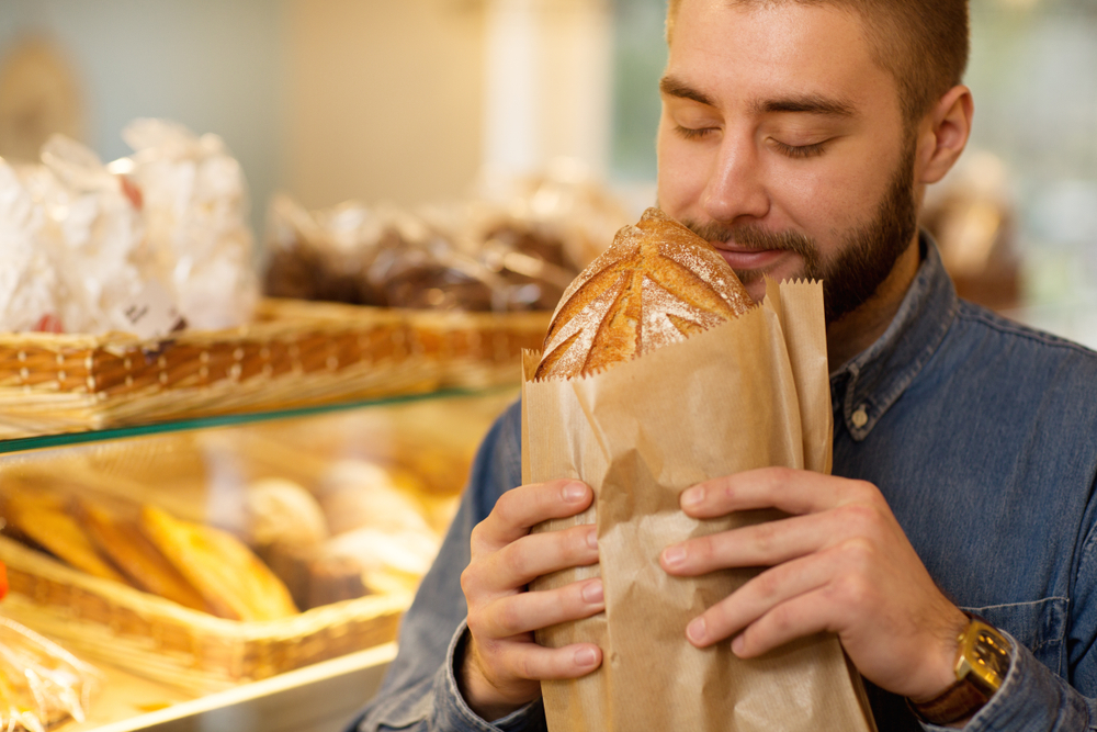 Close up of a young bearded man smelling delicious freshly baked bread at the bakery store(Zoriana Zaitseva)S