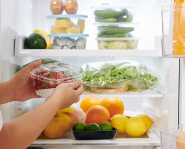 Hands of woman opening fridge door and putting package of fresh ripe strawberries in it