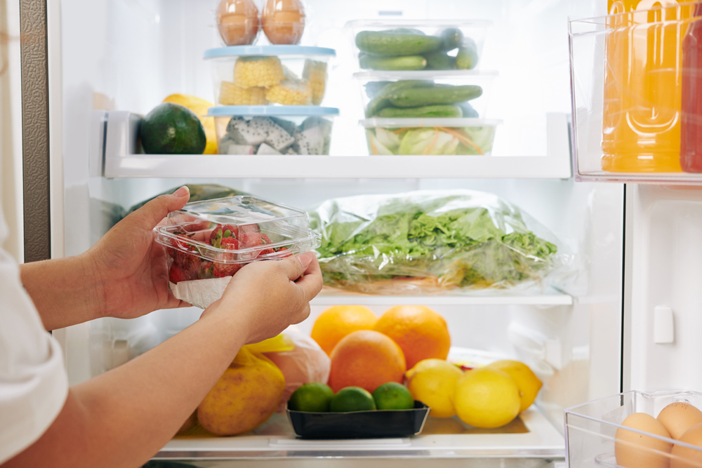 Hands of woman opening fridge door and putting package of fresh ripe strawberries in it