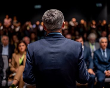 Business man is making a speech in front of a big audience at a conference hall. Speaker giving a talk on corporate business or political conference.