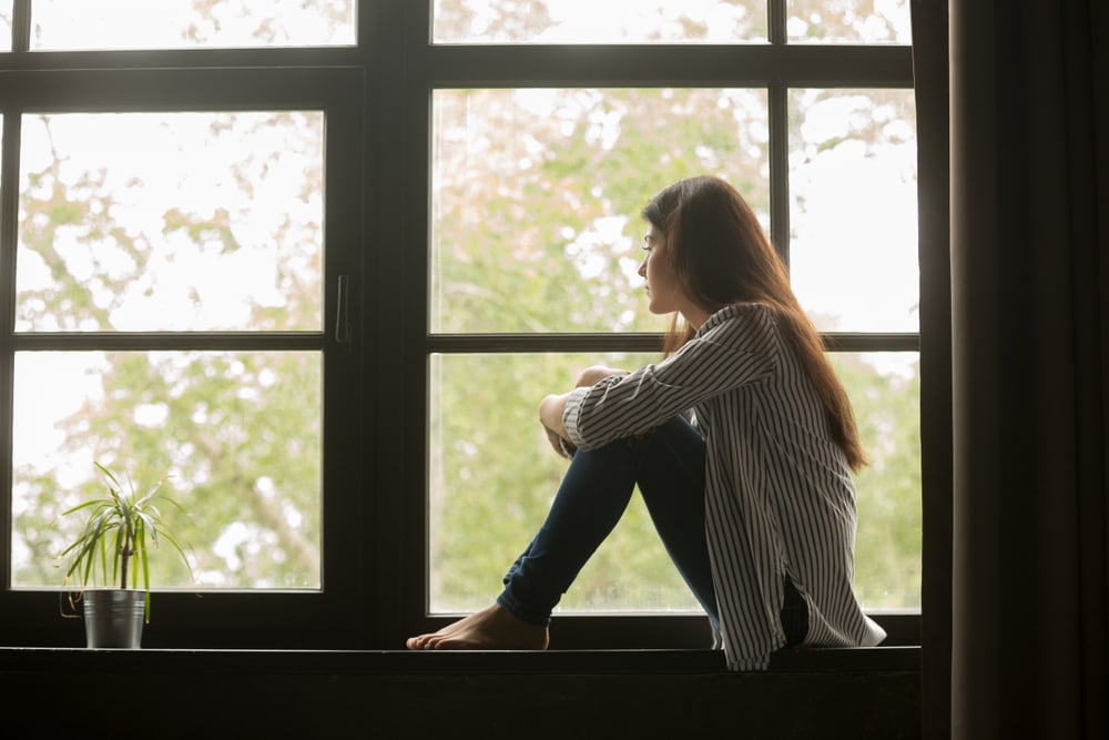 Thoughtful girl sitting on sill embracing knees looking at window, sad depressed teenager spending time alone at home. (fizkes)s