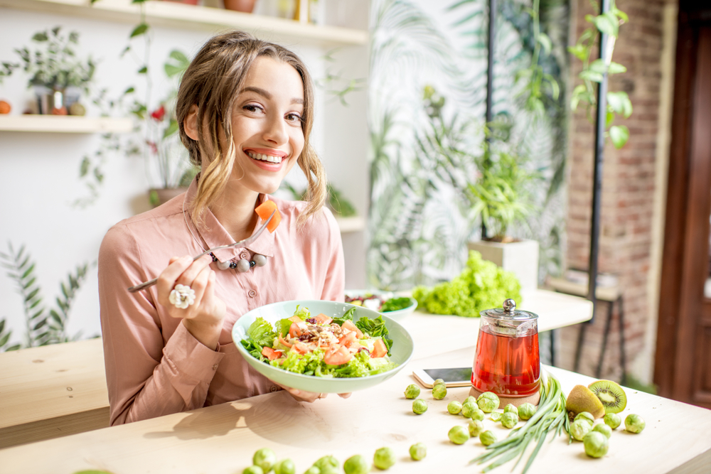 Jovem comendo alimentos saudáveis, sentado no belo interior com flores verdes no fundo (RossHelen) S