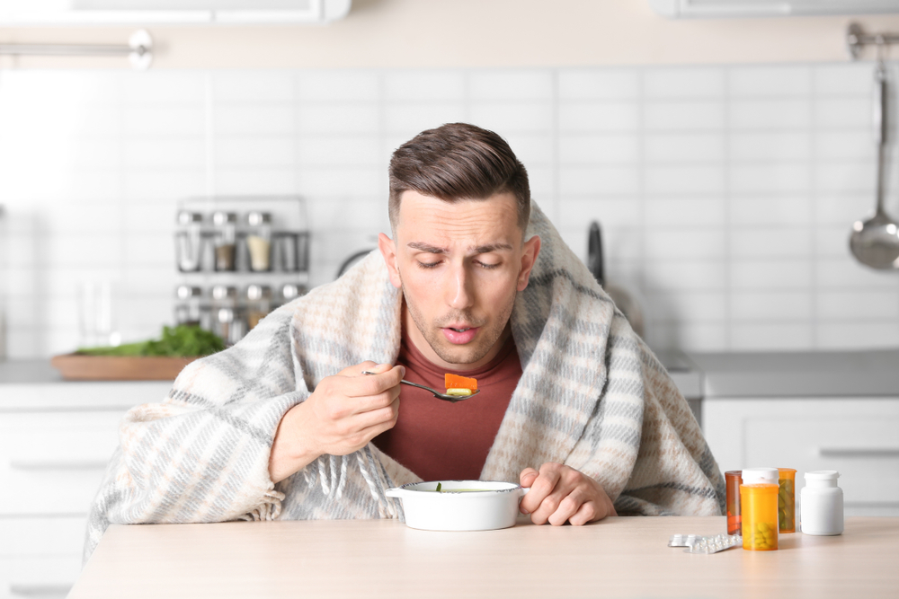 Sick young man eating broth to cure cold at table in kitchen(New Africa)s