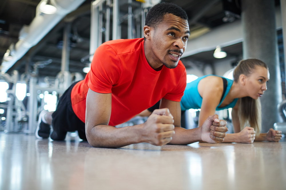 Young athlete in sportswear making effort while doing plank on the floor with his mate near by