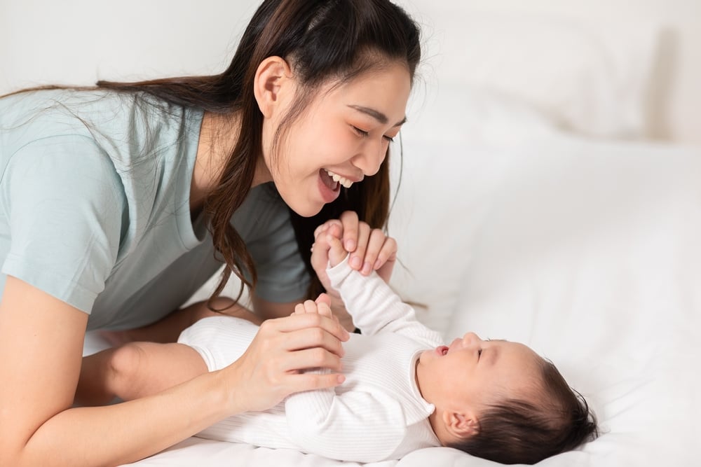 Mamá de Nueva Asia jugando con un adorable recién nacido en la cama sonriendo y feliz en casa.
