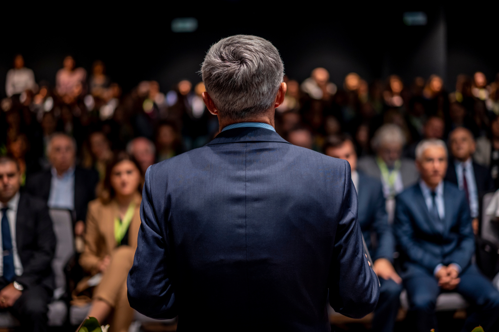 Business man is making a speech in front of a big audience at a conference hall. Speaker giving a talk on corporate business or political conference.